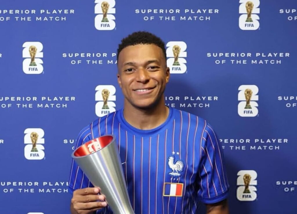 Young man in a blue france jersey holds a tall silver trophy smiling against a fifa backdrop reading superior player of the match